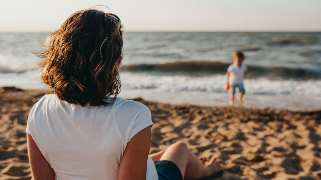En mor sitter svalappet på stranden og ser på barnet sitt leke, vel vtiende at de har full trygghet i WaterCircles Reiseforsirking og Helsetelefonen kundefordel