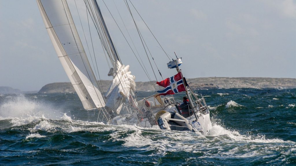 Seilbåt i krevende sjø med det norske kongeflagget/orlogsflagget vaiende i akterenden.